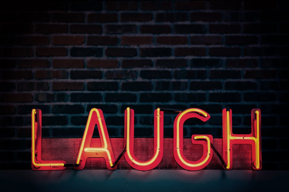 The word ‘laugh’ appears in all caps in a neon sign. It is set in front of a brick wall. The neon lights are an orange hue.