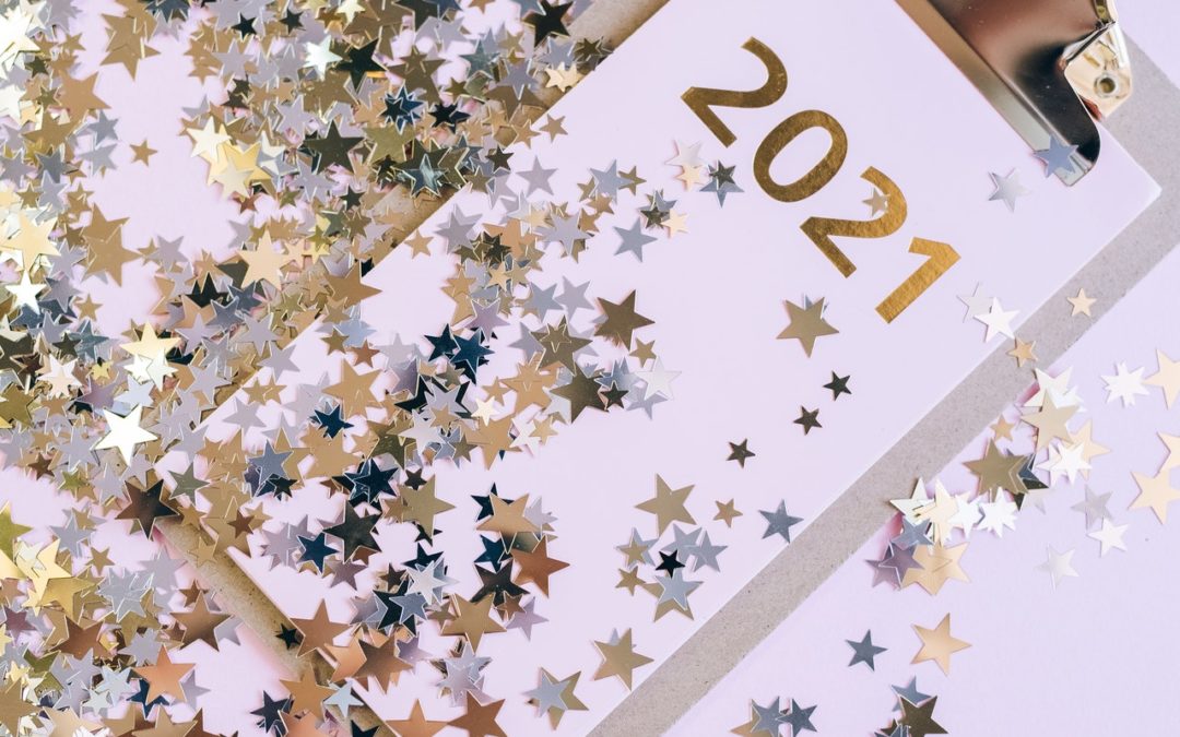 A clipboard with '2021' on it is seen lying at an angle. The clipboard is covered in glitter both on it and surronding it.