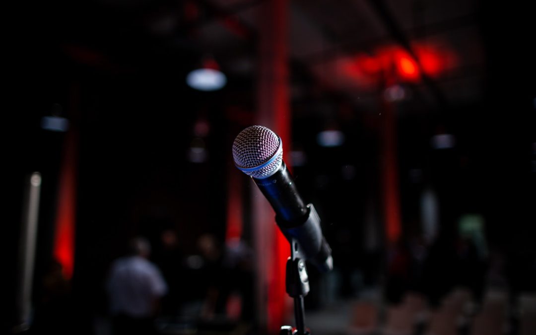 Microphone is seen at close range, on a stand. In the background, out of focus is seating.