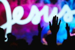 Out-of-focus shot of a neon Jesus sign, with hands in the crowd in the frame.