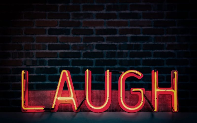 The word ‘laugh’ appears in all caps in a neon sign. It is set in front of a brick wall. The neon lights are an orange hue.